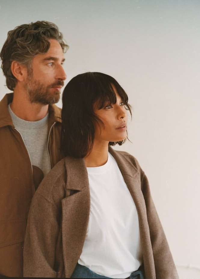 A stylish couple poses indoors. The woman wears a brown coat and the man wears a brown jacket. The background is neutral.