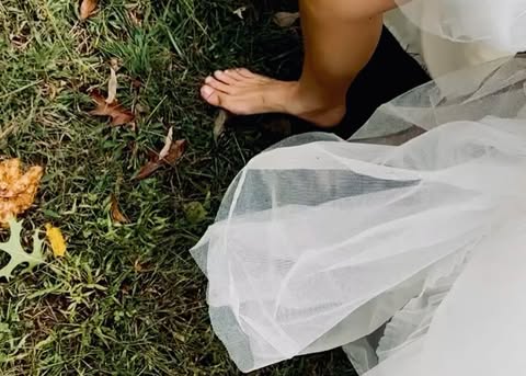 Barefoot woman in a white dress walking on a grassy lawn covered in leaves. The dress is white and flowing, and the grass is green.