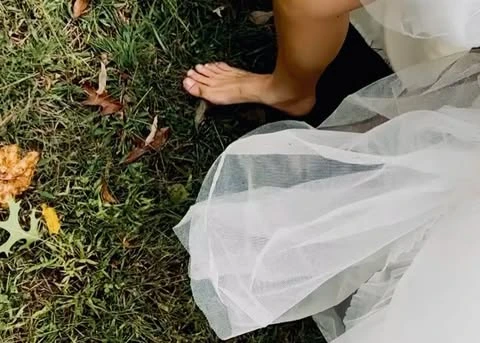 Barefoot woman in a white dress walking on a grassy lawn covered in leaves. The dress is white and flowing, and the grass is green.