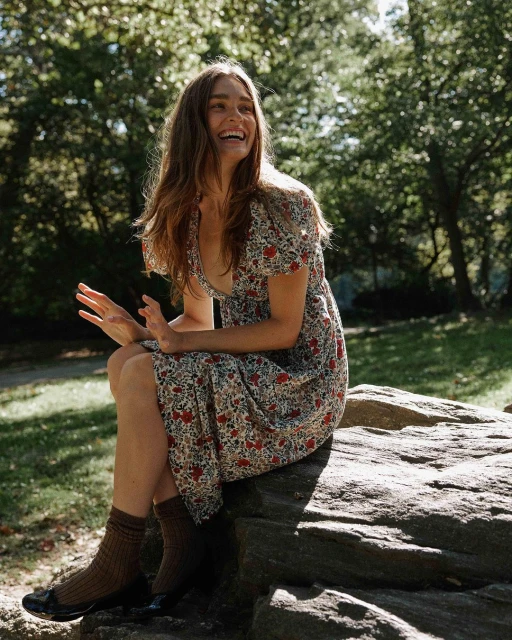 Adult woman in floral dress sits on rock formation in park with trees and foliage in background, smiling and gesturing with her hands.