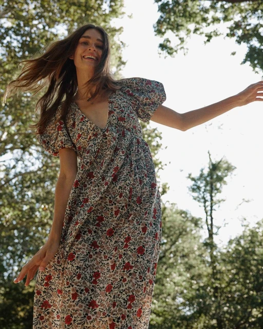A woman in a floral dress smiles with her arms outstretched in a sun-dappled forest environment, viewed from a low angle, with trees visible.