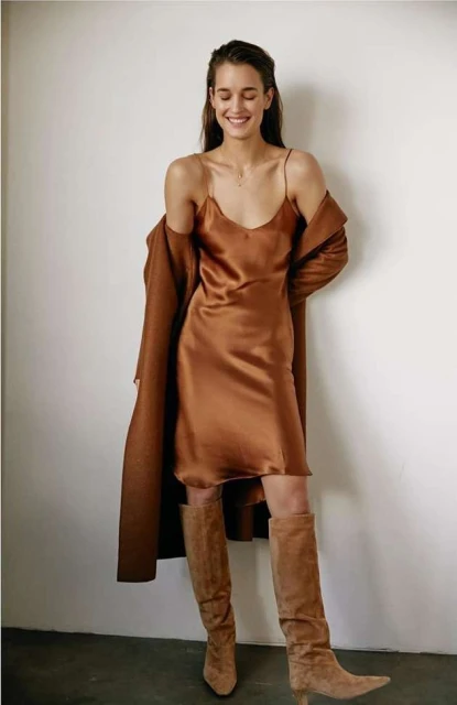 A woman poses indoors wearing a copper slip dress, brown boots, and a matching coat, smiling with hair neatly combed back.