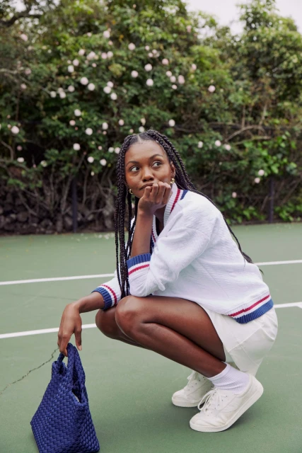 Young woman with braided hair poses in a crouch on a tennis court, wearing a white sweater and skirt, holding a blue woven bag.