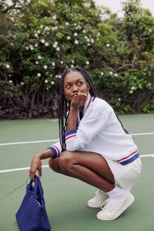 Black woman with braided hair crouching on a tennis court wearing a white sweater, skirt, and sneakers while holding a blue woven handbag.