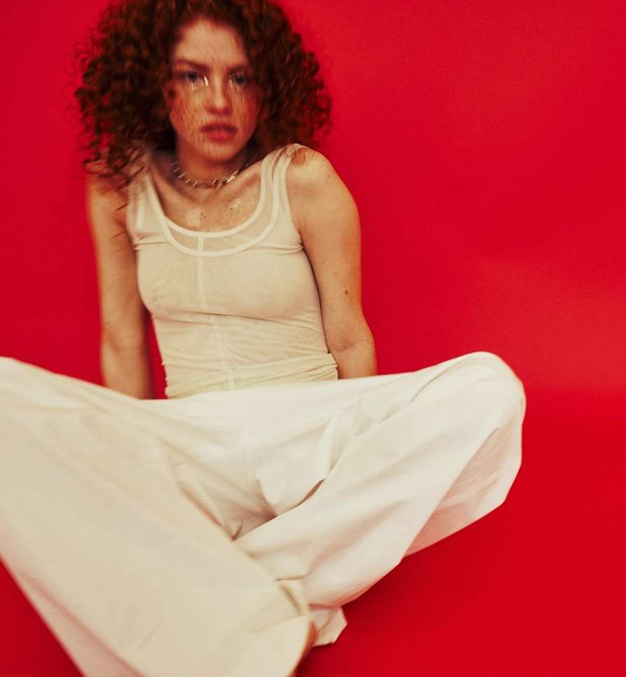 Young woman with red curly hair poses against a red background, wearing a sheer top and white pants while sitting cross-legged.