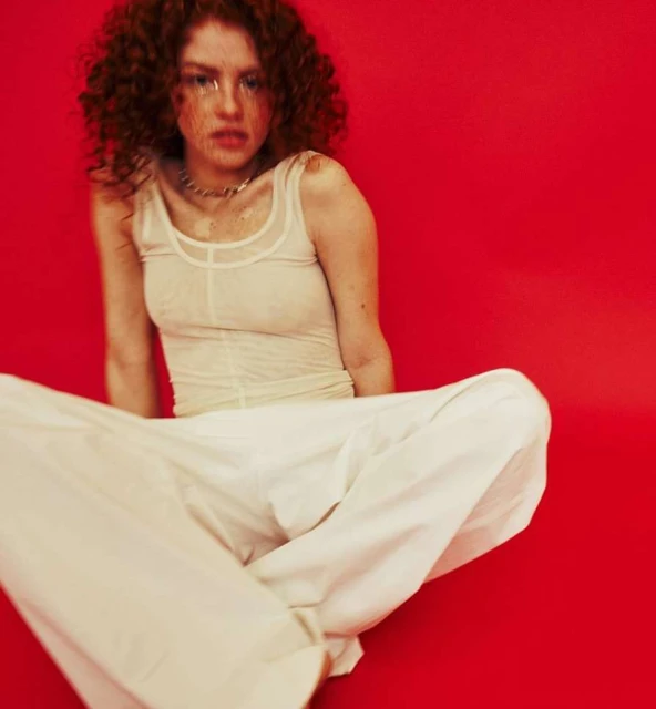 Young woman with red curly hair poses against a red background, wearing a sheer top and white pants while sitting cross-legged.