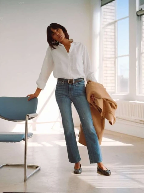 Brunette woman in a white shirt, jeans, and black flats stands in a naturally lit room holding a tan jacket over her shoulder.