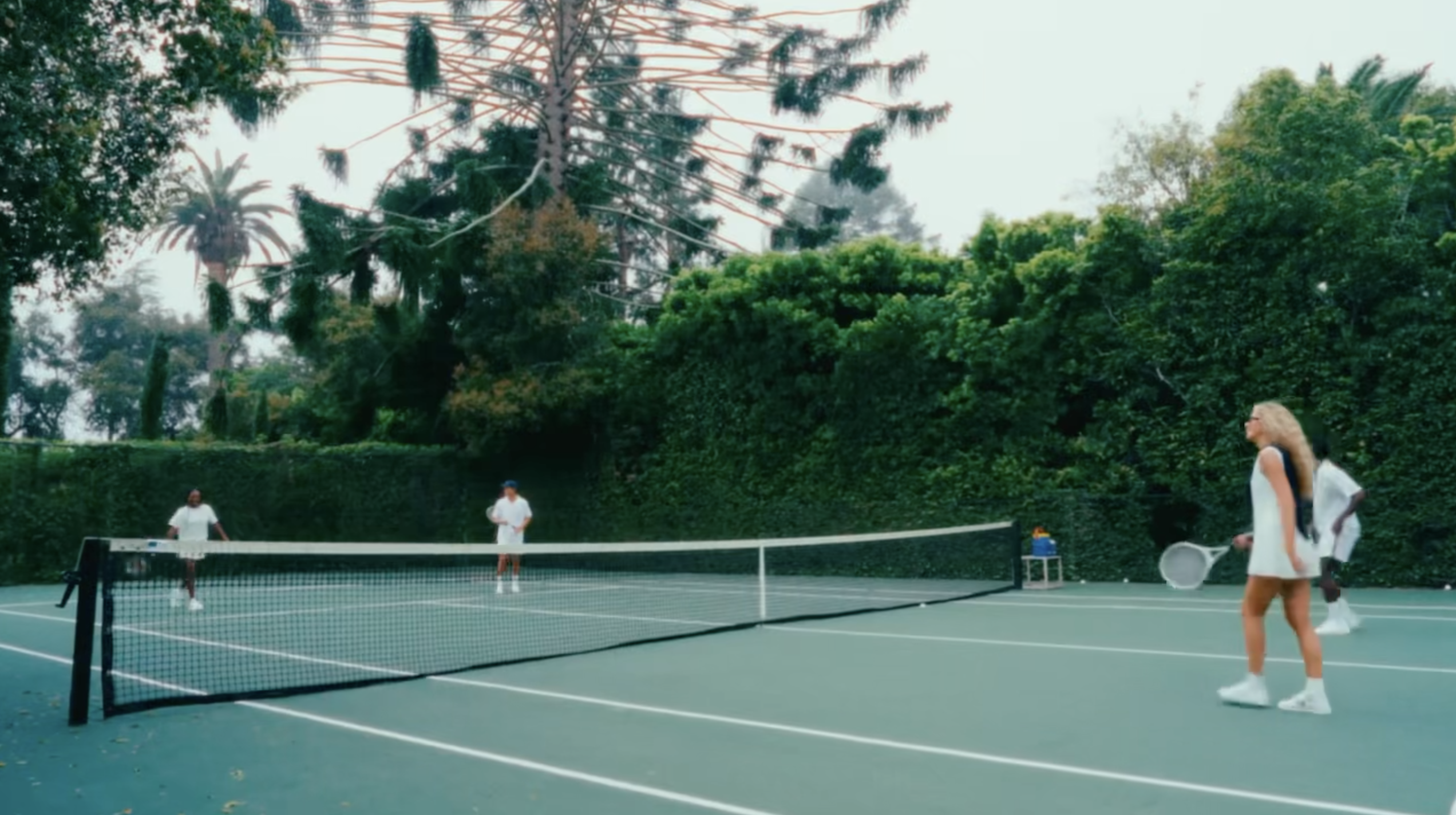 Four adults play tennis on a green court surrounded by lush green trees and bushes on an overcast day with a tennis net.