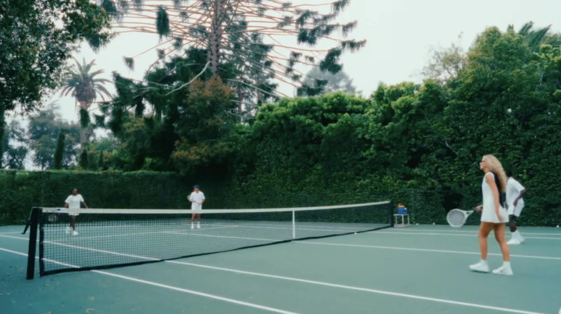 Four adults play tennis on a green court surrounded by lush green trees and bushes on an overcast day with a tennis net.