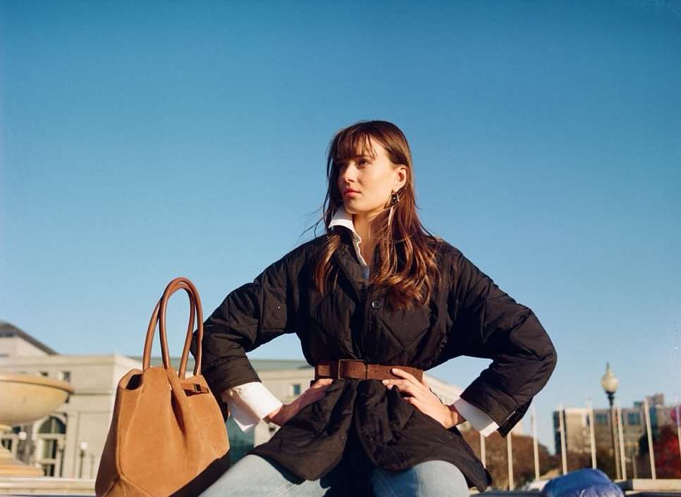 A woman poses outdoors, wearing a black quilted jacket, light blue jeans, and a brown belt, with a tan leather handbag beside her.