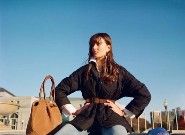 A woman poses outdoors, wearing a black quilted jacket, light blue jeans, and a brown belt, with a tan leather handbag beside her.