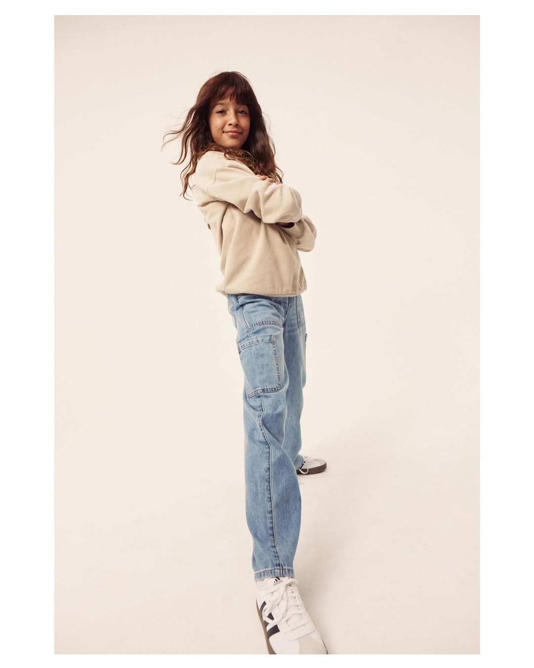 A stylish young girl poses indoors with arms crossed wearing a beige sweater, light blue jeans, and sneakers against a neutral backdrop.