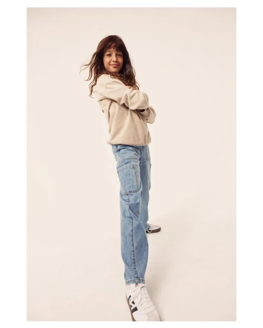 A stylish young girl poses indoors with arms crossed wearing a beige sweater, light blue jeans, and sneakers against a neutral backdrop.