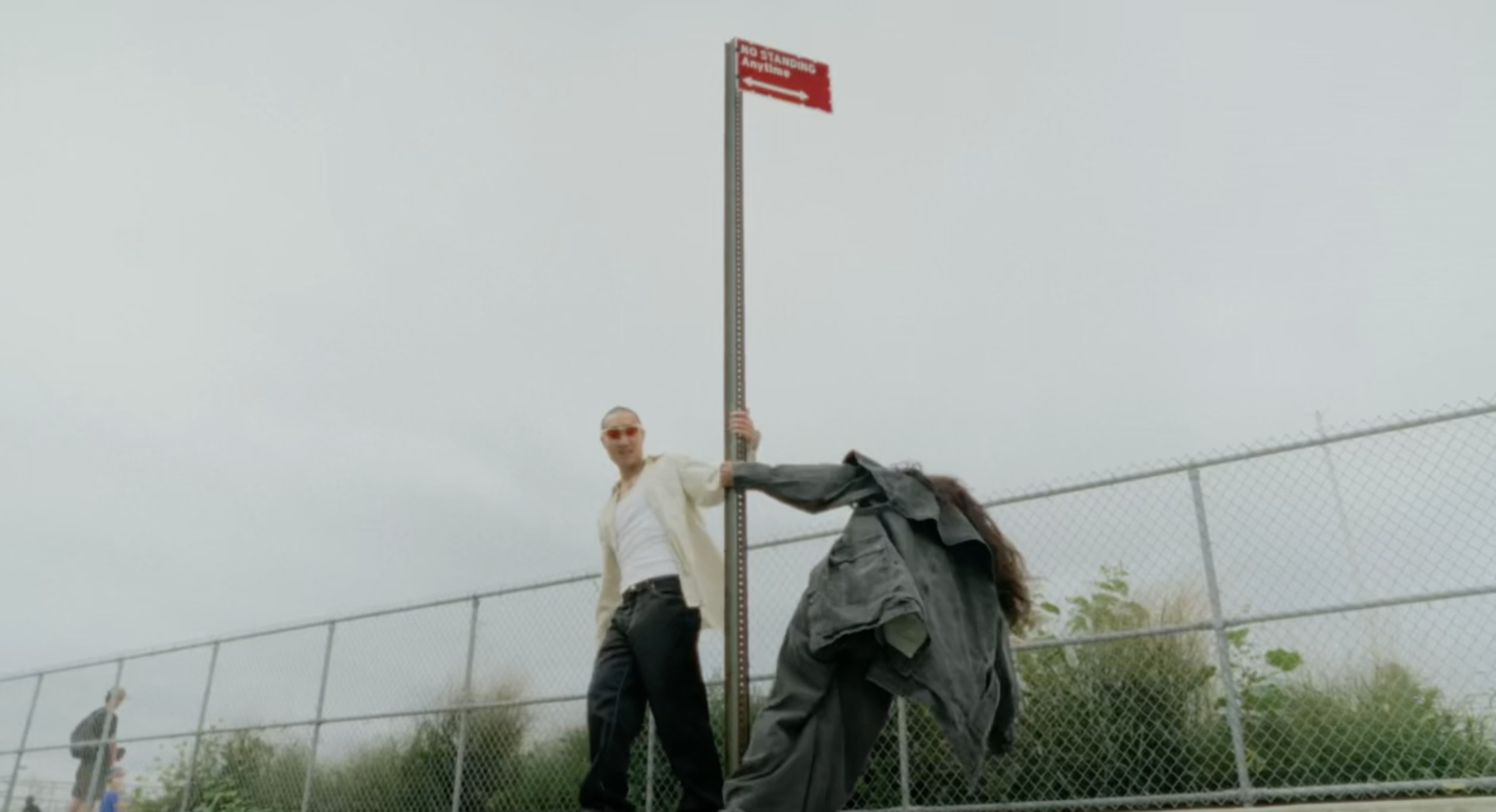 Adult male in an off-white shirt and dark pants poses near a street sign next to a floating denim jacket and pants, outdoors.