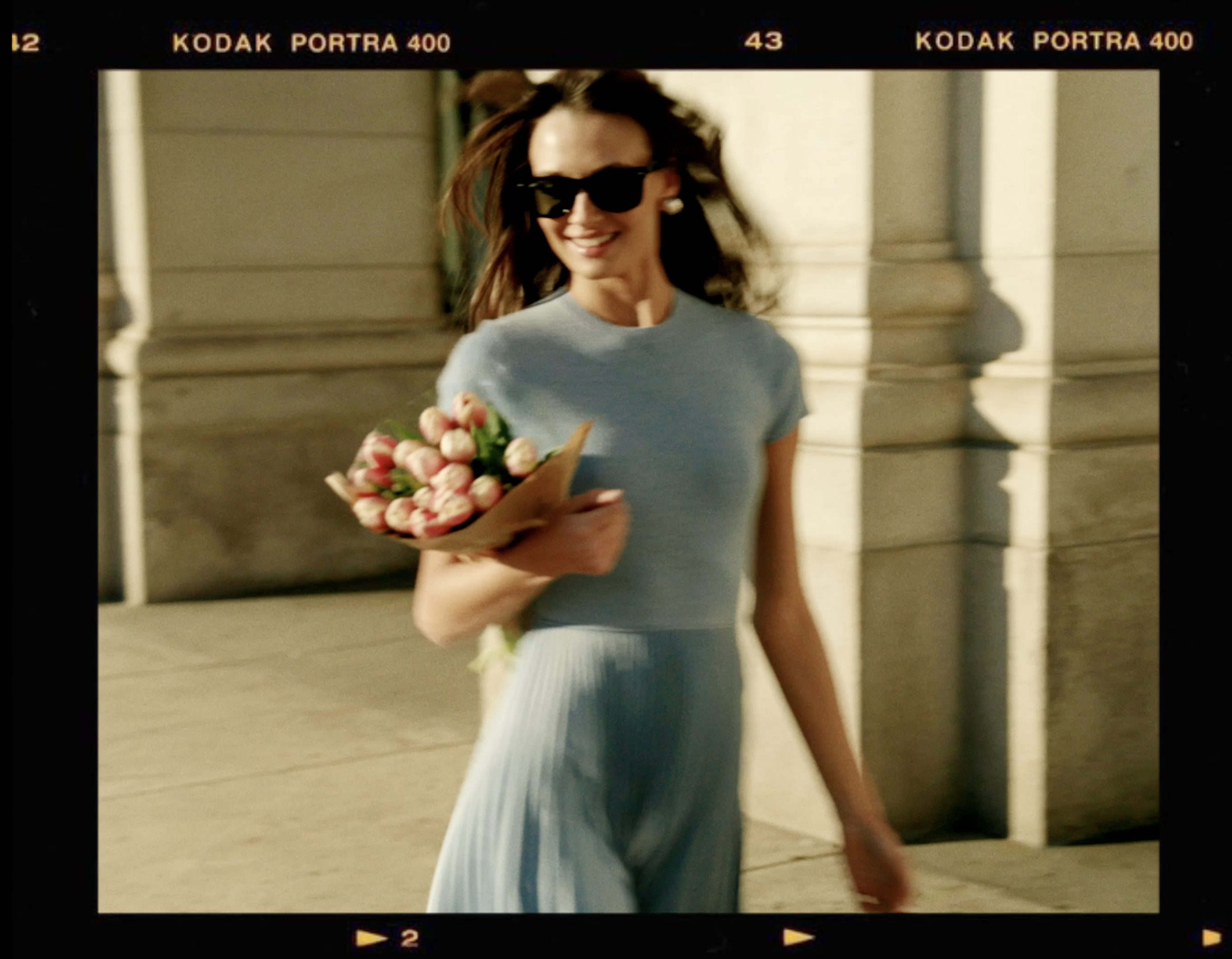 Smiling woman in a blue dress holds a bouquet of pink tulips while walking outside on a sunny day with shadows and film border.