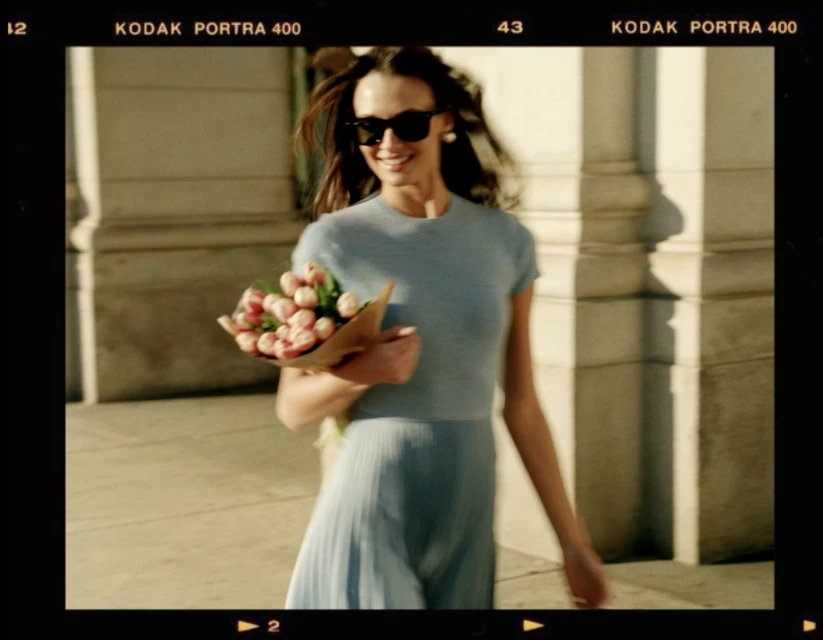 Smiling woman in a blue dress holds a bouquet of pink tulips while walking outside on a sunny day with shadows and film border.