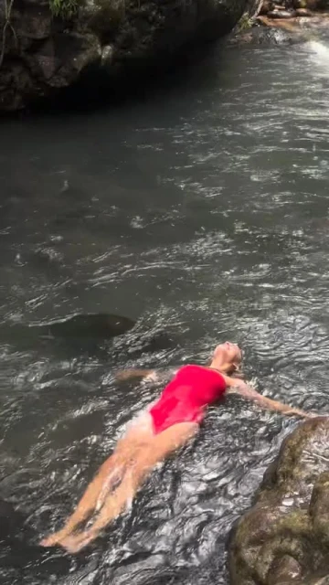 Adult woman in a red swimsuit floats in a river surrounded by rocks and foliage under natural lighting during the daytime.