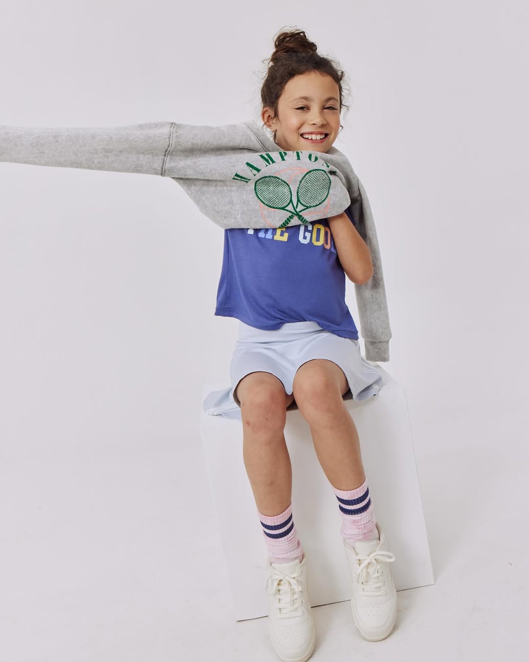 A young girl smiles while sitting on a white cube in a light grey sweatshirt, blue t-shirt, light blue skirt, pink socks, and white shoes.