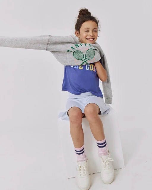 A young girl smiles while sitting on a white cube in a light grey sweatshirt, blue t-shirt, light blue skirt, pink socks, and white shoes.
