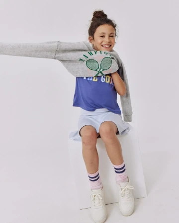 A young girl smiles while sitting on a white cube in a light grey sweatshirt, blue t-shirt, light blue skirt, pink socks, and white shoes.