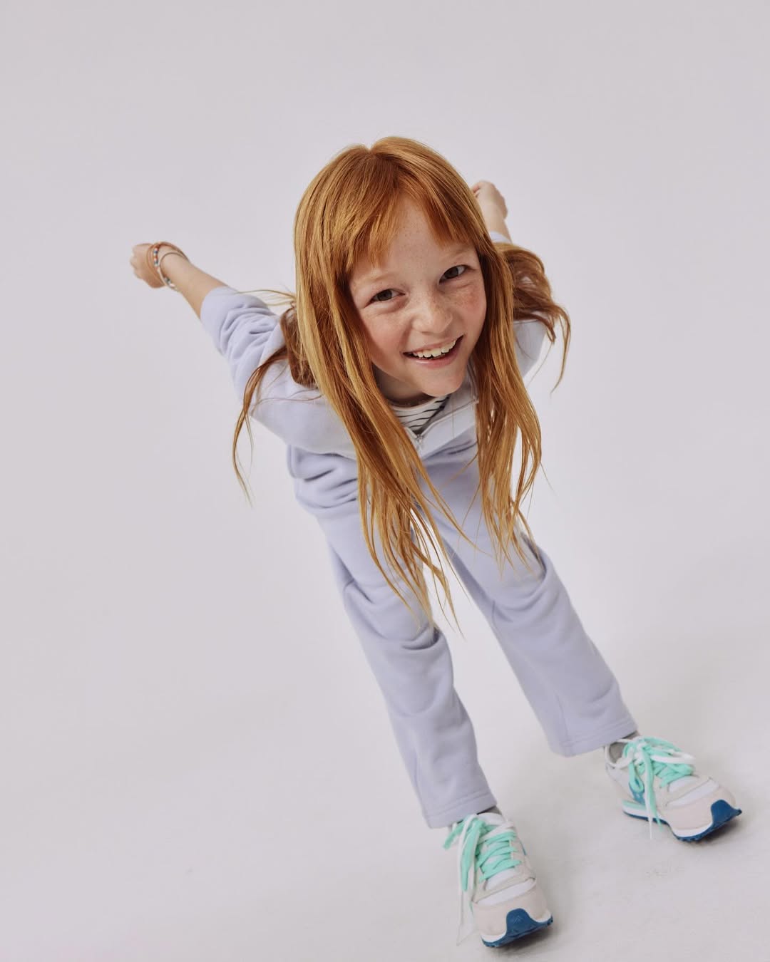 A young red-haired girl in a lavender sweatsuit smiles at the camera from a low angle in a bright white photo studio setting.