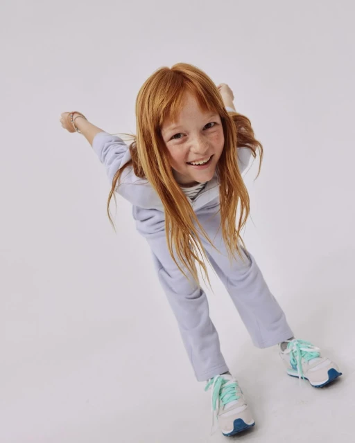 A young red-haired girl in a lavender sweatsuit smiles at the camera from a low angle in a bright white photo studio setting.