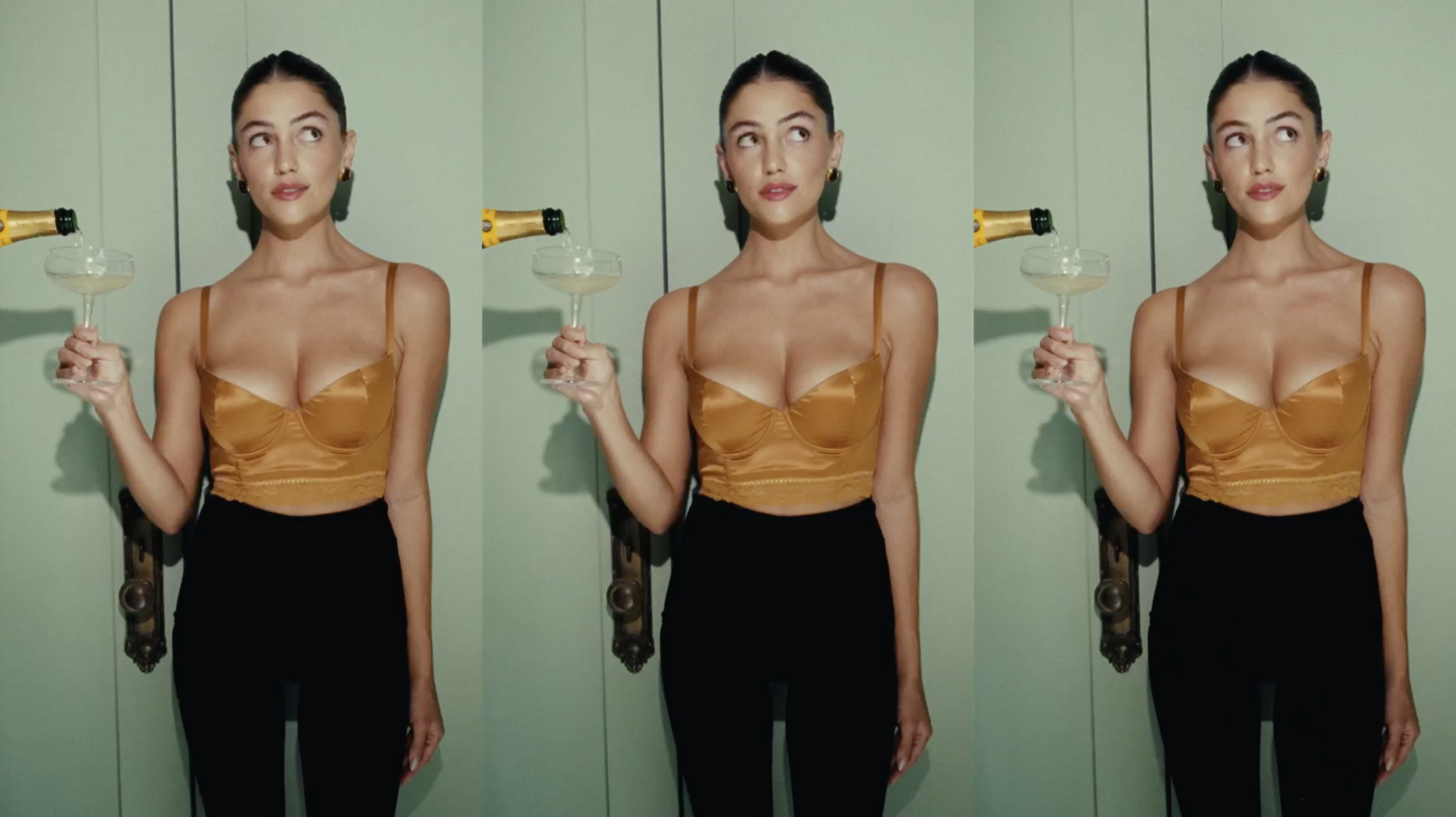 A woman with brown hair in a bronze top and black pants pours champagne into a coupe glass against a light green wall.