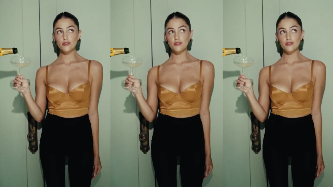 A woman with brown hair in a bronze top and black pants pours champagne into a coupe glass against a light green wall.