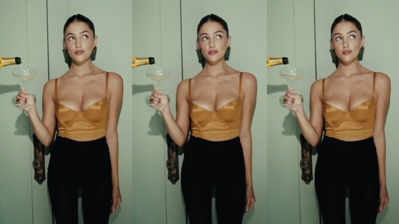 A woman with brown hair in a bronze top and black pants pours champagne into a coupe glass against a light green wall.