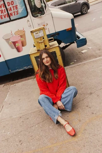 A woman in an orange sweater and jeans sitting on the sidewalk with an ice cream truck and a gray SUV in the background.