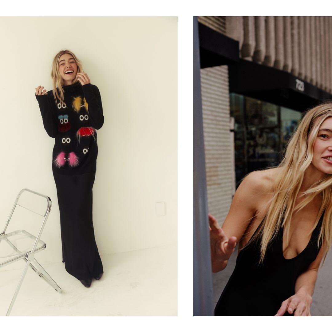 Smiling blond woman in black dress and patterned sweater poses in a brightly lit room, with another woman outside a building.
