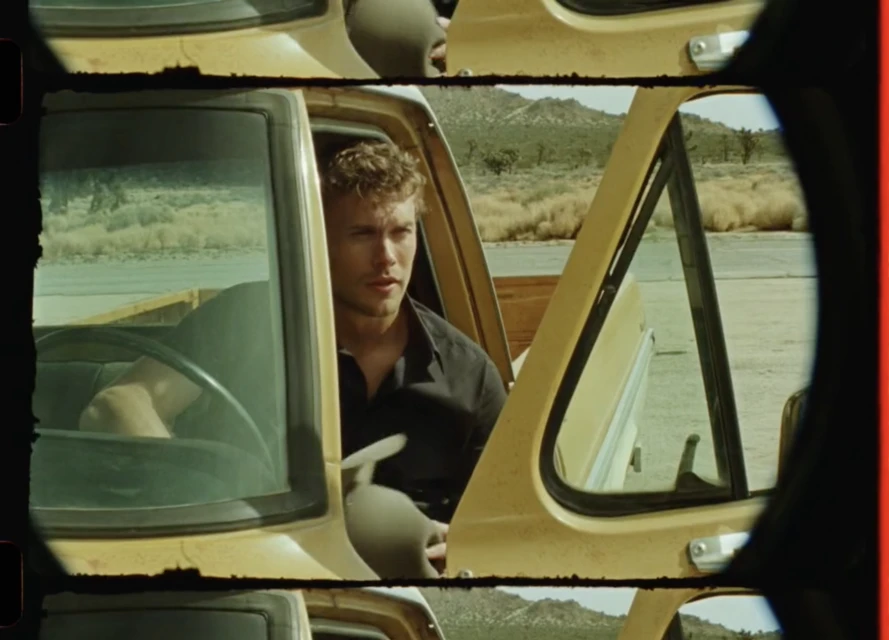 Man in a vintage yellow pickup truck on a desert road, viewed through a film gate. Open door, short curly hair, natural lighting.