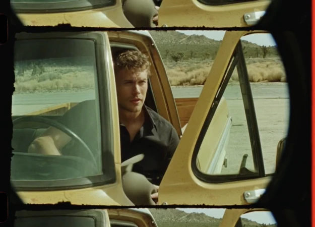 Man in a vintage yellow pickup truck on a desert road, viewed through a film gate. Open door, short curly hair, natural lighting.