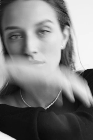 Close-up black and white portrait of a woman wearing a dark top with a thin necklace, captured with motion blur across her face.