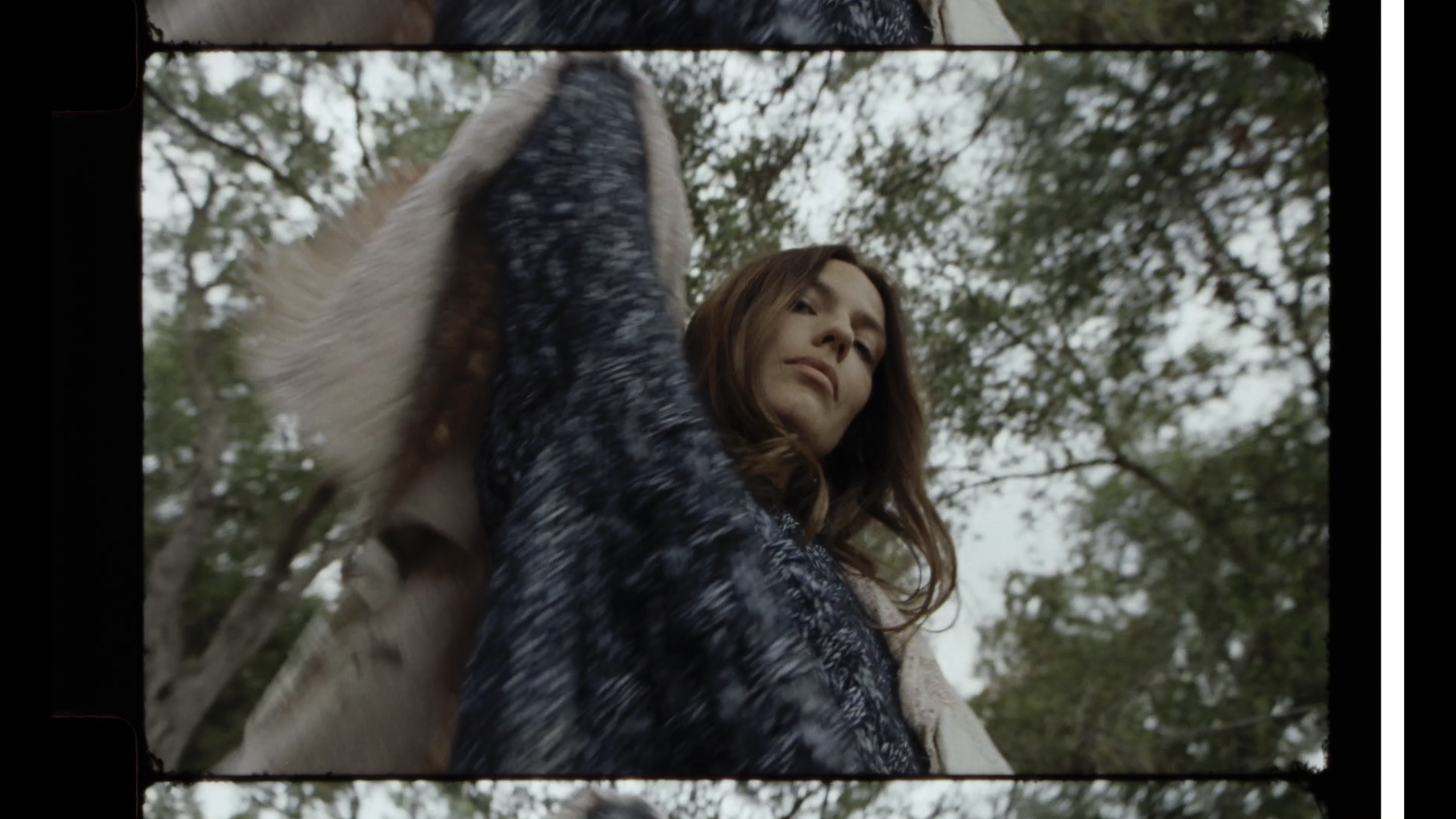 An adult female in a faux fur coat glances downward in a low-angle outdoor shot framed by trees and branches under overcast light.