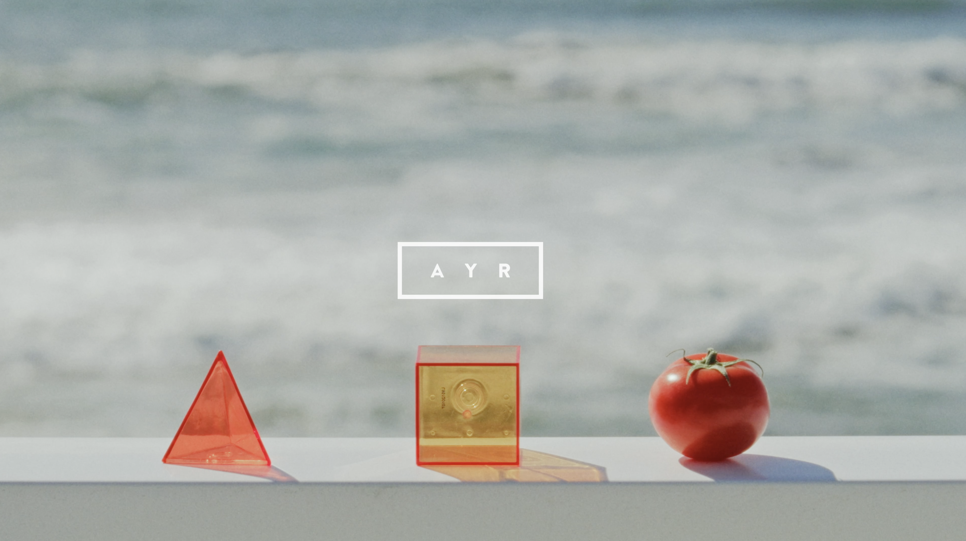 Still life with red translucent geometric objects and a tomato on a white ledge with an ocean background and AYR logo.