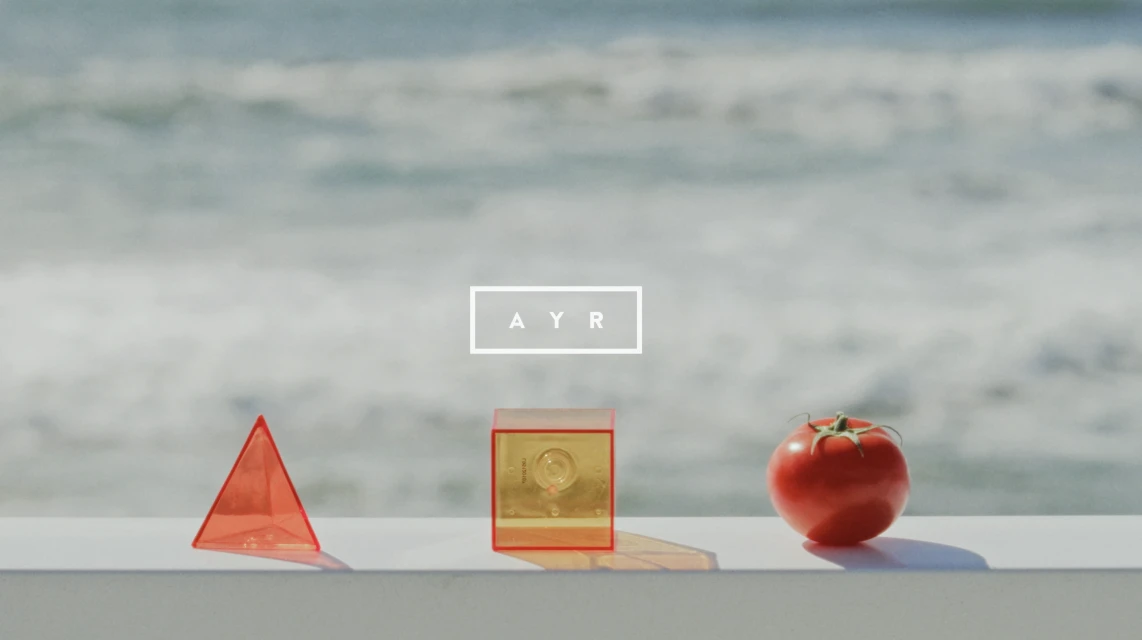 Still life with red translucent geometric objects and a tomato on a white ledge with an ocean background and AYR logo.