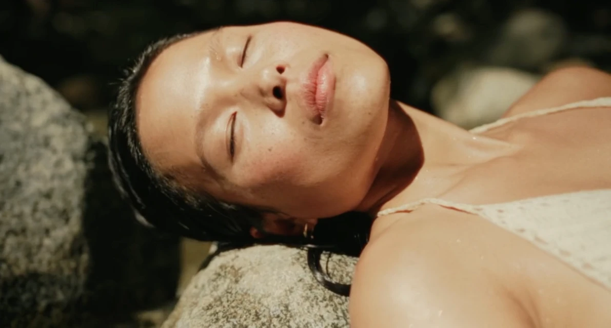 Portrait of a relaxed Asian woman lying on rocks, eyes closed, with a blurred background of foliage and warm, natural light.