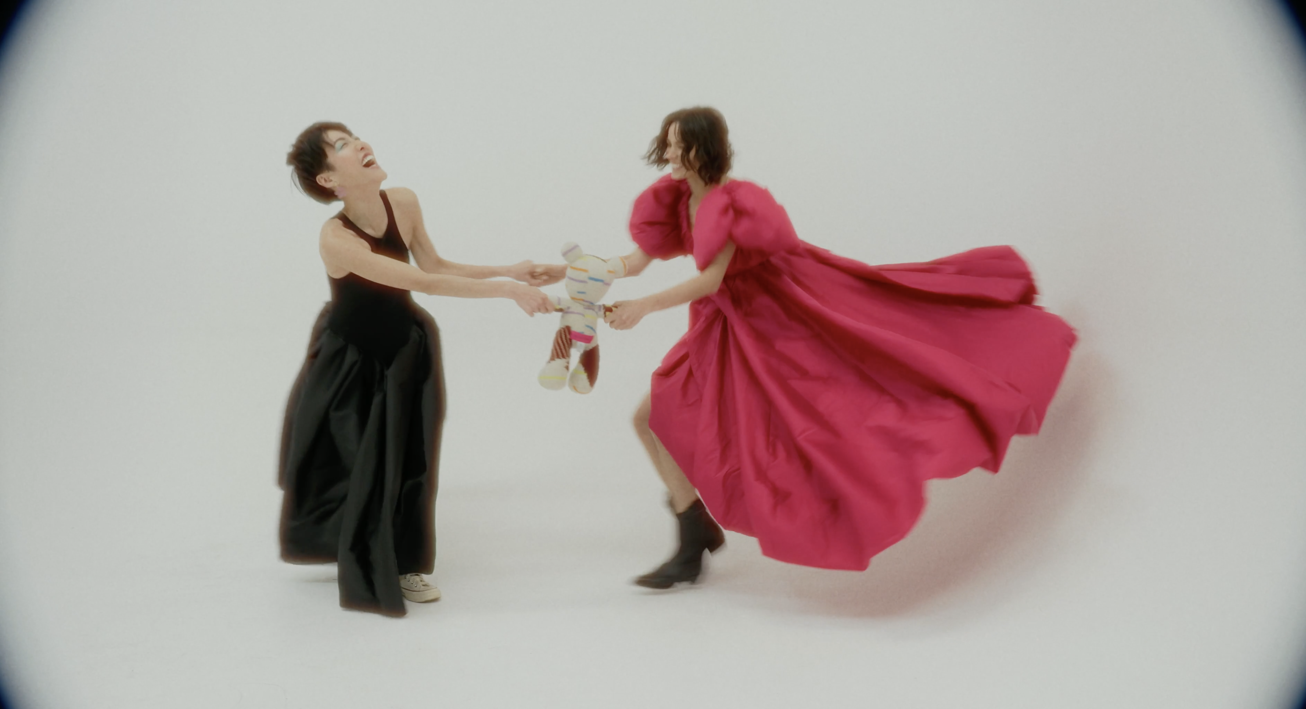 Two women in bright outfits playfully tug at a striped stuffed toy in a bright photo studio, laughing and wearing fashionable clothing.