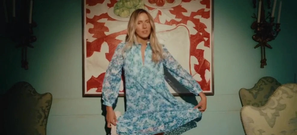Blonde woman in blue floral dress stands in front of a painting in an interior room with green walls and antique furnishings.