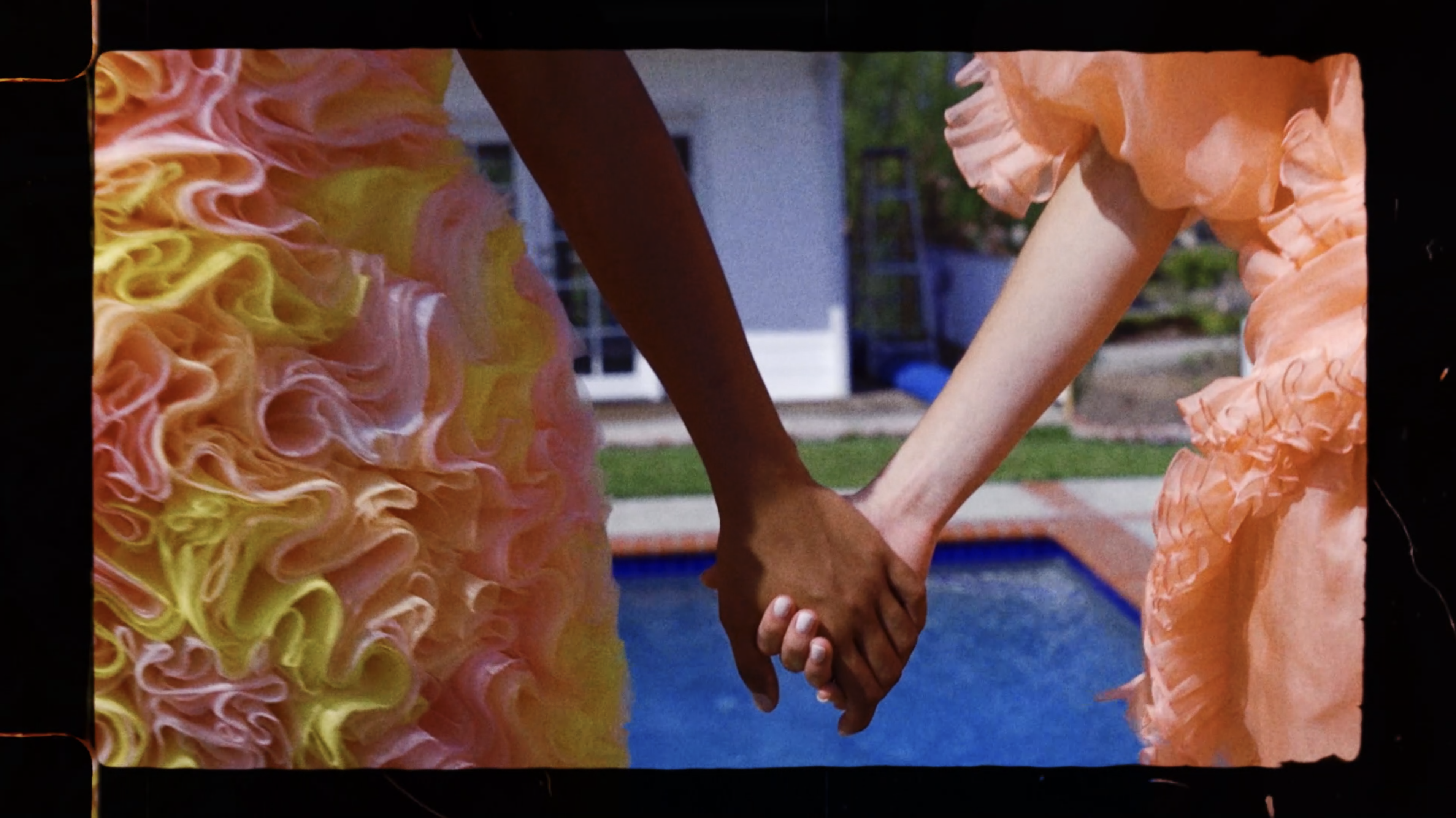 Two people hold hands in front of a pool, one wears a ruffled yellow dress, the other a ruffled orange dress. A white house is visible.
