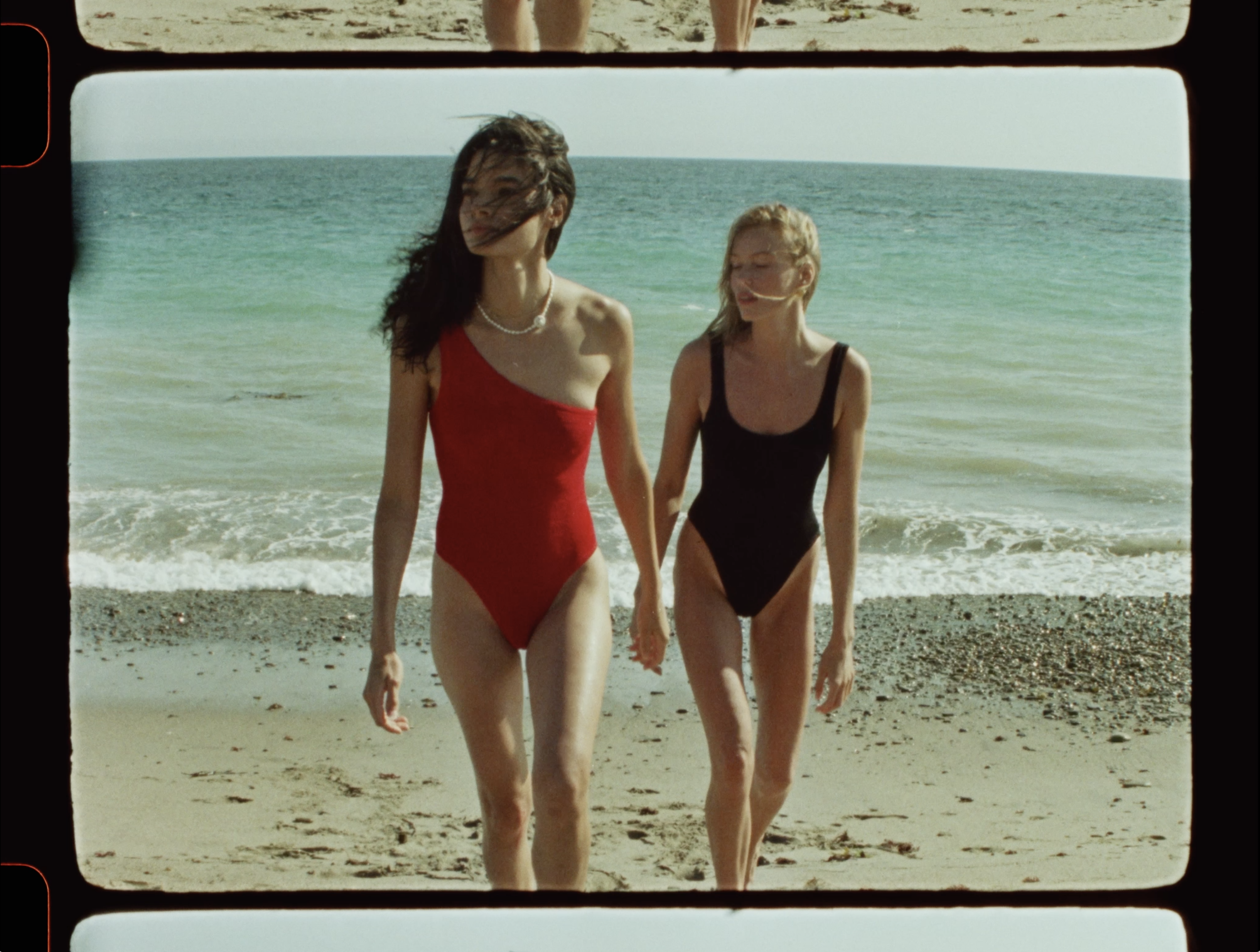 Two women in swimsuits walk on a beach with light-colored sand, against a backdrop of a blue-green ocean and sky on a sunny day.