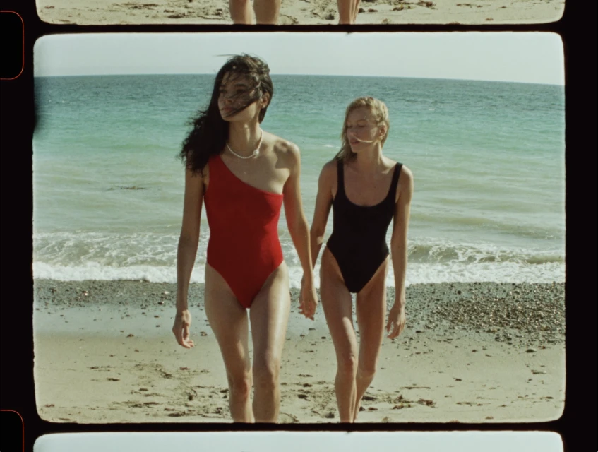 Two women in swimsuits walk on a beach with light-colored sand, against a backdrop of a blue-green ocean and sky on a sunny day.