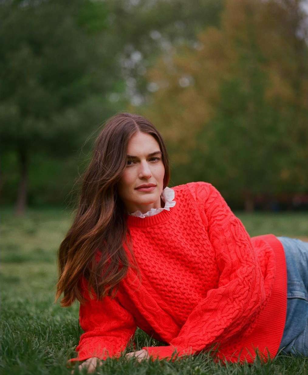 Woman with long brown hair lying on green grass wearing a bright red cable-knit sweater with a white lace collar
