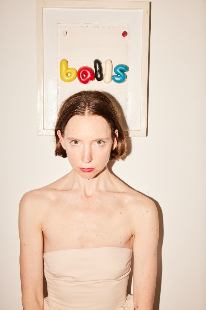 Woman in nude strapless top stands beneath a framed artwork spelling out 'balls' in colorful bubble letters, portrait framing indoors.