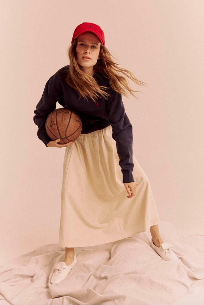 Female model in navy sweatshirt, cap, and skirt holding a basketball in a studio environment with a light pink background.