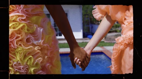 Two people holding hands outdoors by a swimming pool wearing ruffled dresses, captured in vintage 16mm film format.