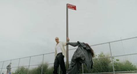 Two people posing near a tall street sign post with a chain-link fence and overcast sky in the background