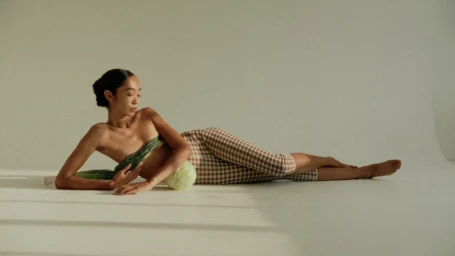 Model lying on side wearing brown gingham trousers and green top against a seamless white studio background