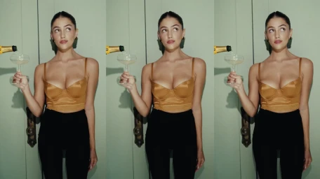 Woman in a gold satin bustier top holding a champagne glass while a bottle pours wine against a green wall